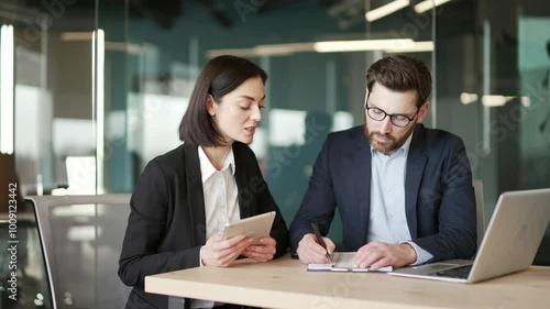Two co-workers are busy with paper work, discuss work issues while sitting at workplace in business office. Male and female worker reviewing documents, filling out form, talking about financial report