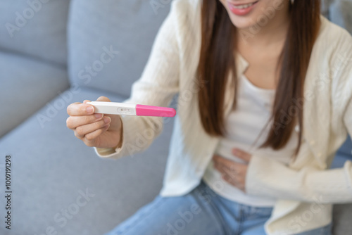 Happy thankful asian young woman hand holding pregnancy test after result with positive, surprised glad girl sitting on couch living room at home. Pregnant of ovulation fertility, motherhood concept.