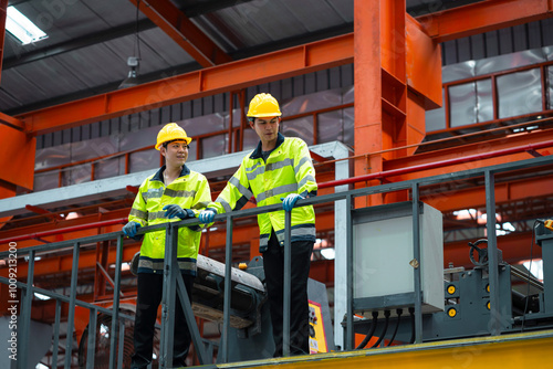 Two men in yellow and black safety gear stand on a platform in a factory. They are looking at something on the ground