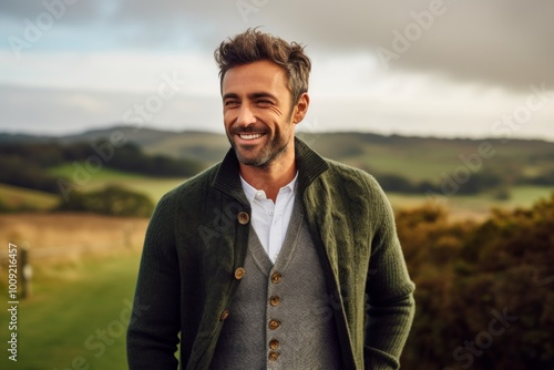 Portrait of a smiling man in his 30s wearing a chic cardigan in front of quiet countryside landscape