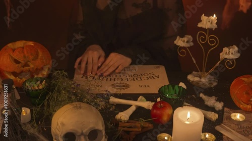 A witch in black clothing conjures a Ouija board while sitting in a scary kitchen with pumpkins and potions. photo