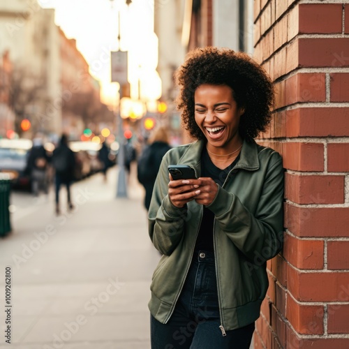 A joyful woman in casual streetwear leans against a brick wall, laughing as she reads her phone, with a city street and warm afternoon light behind her.