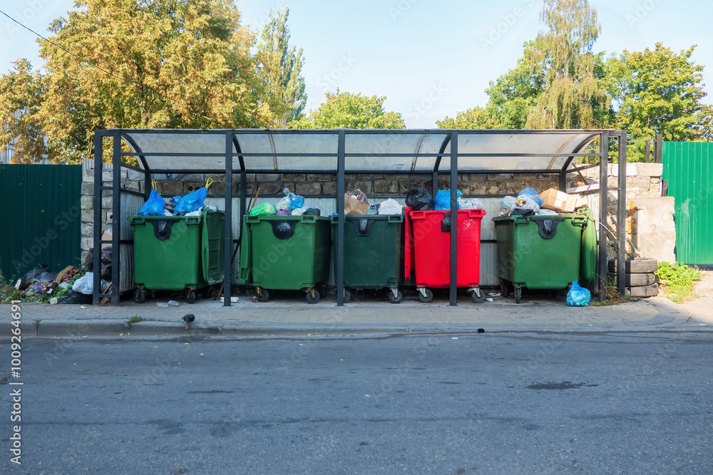 A fragment of a street with multi-colored garbage containers on a special roofed area. In the foreground there is an asphalt road, in the background there are tree branches.