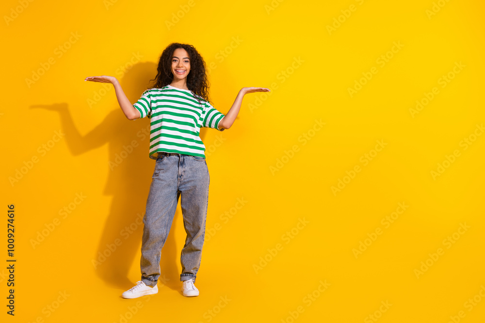 Full body portrait of nice young girl arms hold empty space wear striped t-shirt isolated on yellow color background Full body portrait of nice young girl arms hold empty space wear striped t-shirt isolated on yellow color background