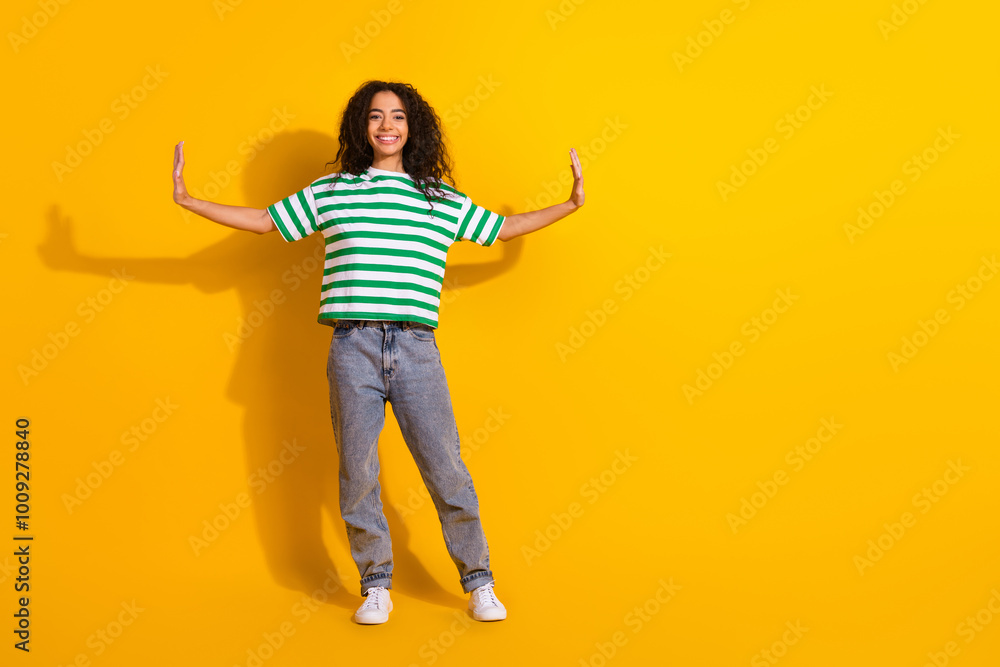 Full size photo of pretty young girl arms push empty space wear striped t-shirt isolated on yellow color background Full size photo of pretty young girl arms push empty space wear striped t-shirt isolated on yellow color background