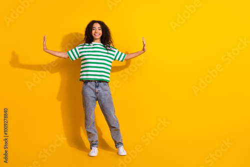 Full size photo of pretty young girl arms push empty space wear striped t-shirt isolated on yellow color background