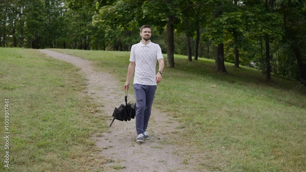 Caucasian man walks through park with folded umbrella in hand on cloudy day after rain. Contented guy strolls along path, improving health and breathing fresh air. Caucasian man walks through park with folded umbrella in hand on cloudy day after rain. Contented guy strolls along path, improving health and breathing fresh air.