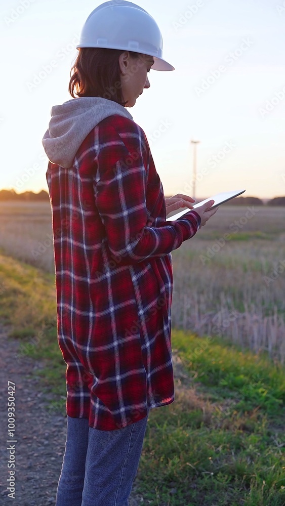 Woman engineer wearing a white protective helmet is taking notes with a clipboard in a field with wind turbines, as the sun sets. Clean energy and engineering concept Woman engineer wearing a white protective helmet is taking notes with a clipboard in a field with wind turbines, as the sun sets. Clean energy and engineering concept