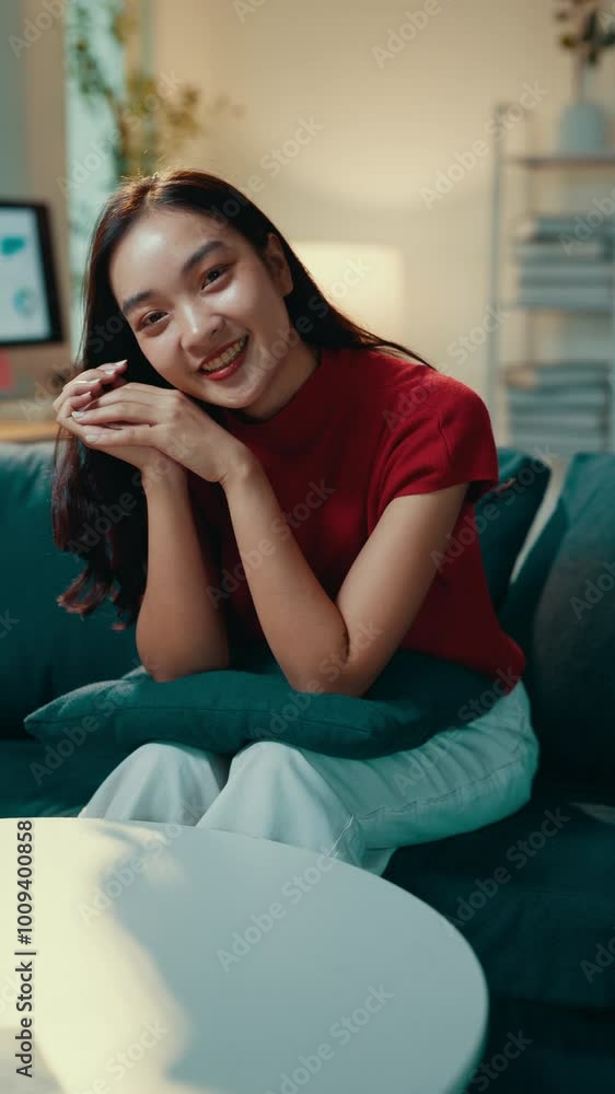 Young woman in a red shirt sits comfortably on a couch, smiling brightly at the camera. Her cheerful expression radiates happiness and confidence in a cozy home setting Young woman in a red shirt sits comfortably on a couch, smiling brightly at the camera. Her cheerful expression radiates happiness and confidence in a cozy home setting