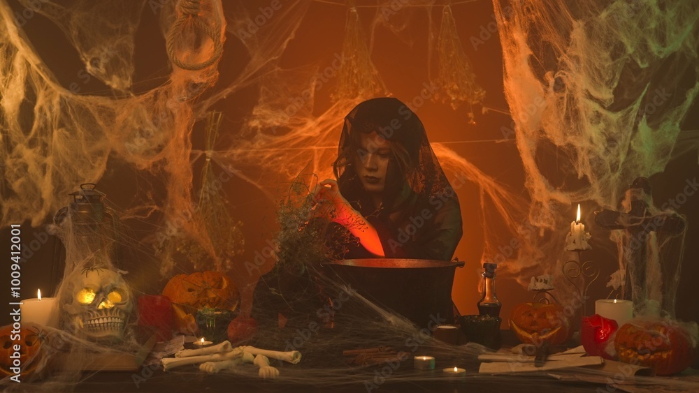 A witch in black clothes is cooking a potion in a scary kitchen with pumpkins and a human skull and bones. A witch in black clothes is cooking a potion in a scary kitchen with pumpkins and a human skull and bones.
