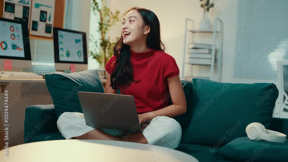 Young asian woman, comfortably dressed, is sitting on a sofa in her home office, working on her laptop. She is smiling and seems to be enjoying her work Young asian woman, comfortably dressed, is sitting on a sofa in her home office, working on her laptop. She is smiling and seems to be enjoying her work