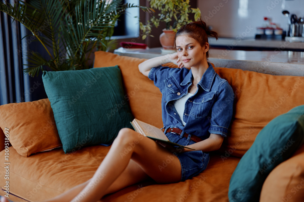 Obraz premium Relaxed woman sitting on orange couch with crossed legs and holding a book in hands by the window
