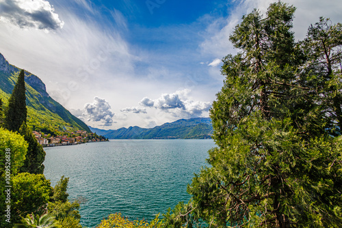 A stunning view of Lake Como with vibrant greenery and palm trees in the foreground. The crystal-clear water and surrounding mountains bask in the bright blue sky and fluffy clouds