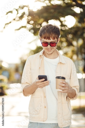 Smiling Young Man Texting on Smartphone and Holding Coffee in Morning City Street