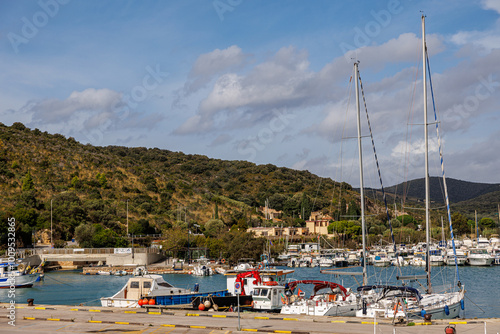 Scenic view of the marina in Talamone, Tuscany, with sailboats docked against a backdrop of lush hills under a clear sky. A picturesque Italian seaside town