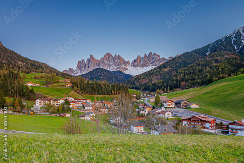 A picturesque Alpine village nestled in green hills with the stunning backdrop of the snow-capped Dolomite mountains. The serene landscape showcases beautiful homes and lush countryside under a clear 