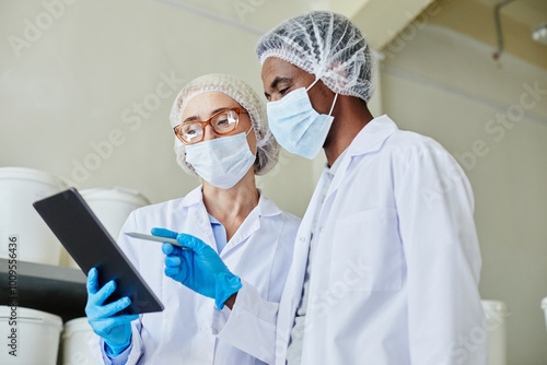 Medium shot of female process technician wearing glasses and mask using digital tablet with African American male coworker while taking inventory of products at pharmaceutical plant, copy space