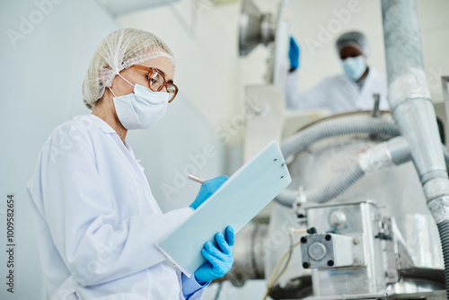 Side view of female technician in protective mask taking notes on notepad while inspecting manufacturing machine on shop floor of pharmaceutical factory, copy space