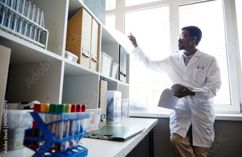 Medium full shot of African American male scientist reaching out to take document from cabinet shelf doing paperwork standing against window in laboratory of scientific research service, copy space