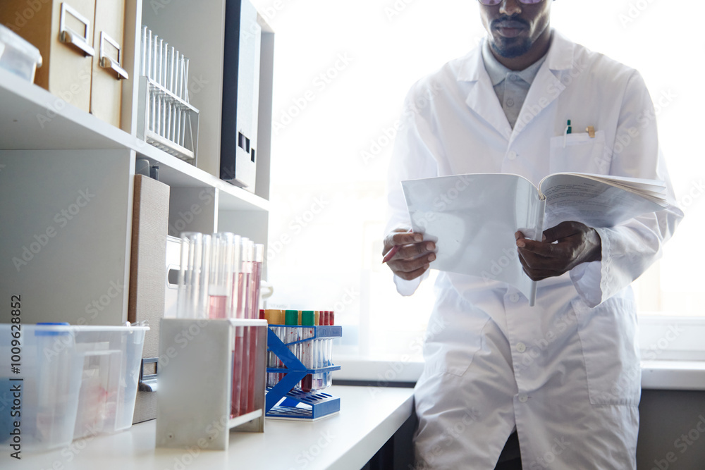 Fototapeta premium Cropped shot of African American male lab chemist reading book next to window performing scientific research in pharmaceutical laboratory, copy space