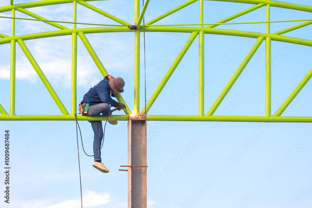 Obraz premium Welder is welding metal of industrial warehouse building structure against blue sky in construction site area