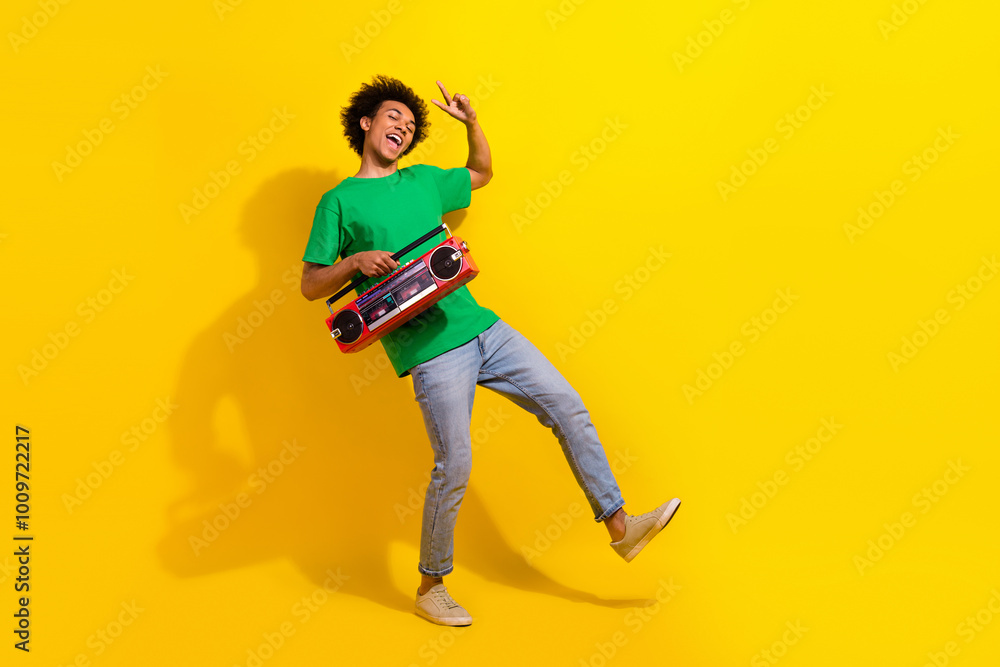 Full length photo of handsome good mood guy wear green t-shirt showing v-sign enjoy boom box songs emtpy space isolated yellow color background Full length photo of handsome good mood guy wear green t-shirt showing v-sign enjoy boom box songs emtpy space isolated yellow color background