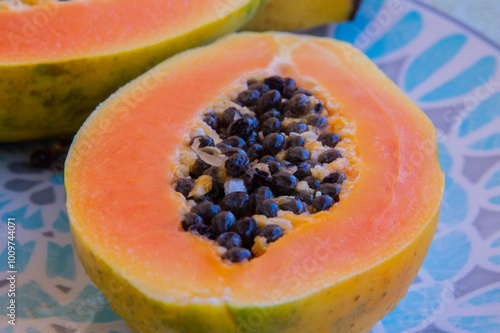 close-up: big orange hue ripe fruit of papaya cut in two with  large central cavity are attached numerous black seeds on a turquois blue plate with rhombus pattern