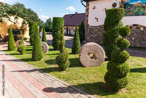 Well-kept garden with a variety of topiary shrubs and a decorative stone mill wheel. With a brick pathway leading into the center of the garden. The trees and shrubs have been carefully shaped. photo