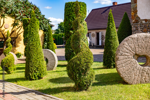 Well-kept garden with a variety of topiary shrubs and a decorative stone mill wheel. With a brick pathway leading into the center of the garden. The trees and shrubs have been carefully shaped. photo