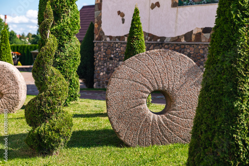 Well-kept garden with a variety of topiary shrubs and a decorative stone mill wheel. With a brick pathway leading into the center of the garden. The trees and shrubs have been carefully shaped. photo