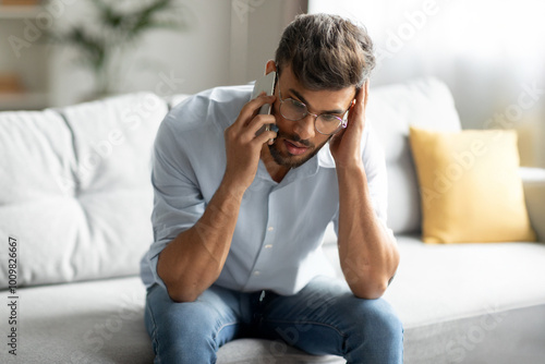 Confused indian guy talking on cellphone and touching head, having unpleasant mobile conversation at home, sitting on sofa in living room interior