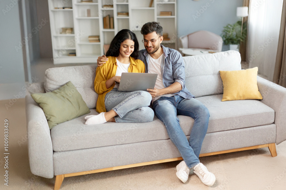 Planning vacation. Happy young indian couple searching tour online, using laptop computer, sitting on couch in cozy living room interior at home, free space Planning vacation. Happy young indian couple searching tour online, using laptop computer, sitting on couch in cozy living room interior at home, free space