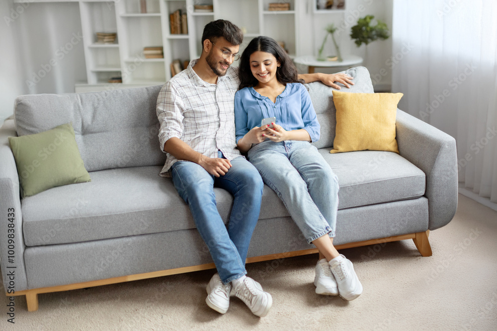 Indian couple using cellphone together, sitting in casual home interior on sofa, surfing internet or chatting with friends. Modern, digitally connected lifestyle Indian couple using cellphone together, sitting in casual home interior on sofa, surfing internet or chatting with friends. Modern, digitally connected lifestyle