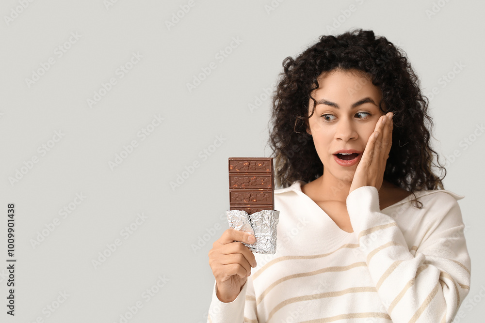 Shocked young African-American woman with sweet chocolate bar on grey background Shocked young African-American woman with sweet chocolate bar on grey background