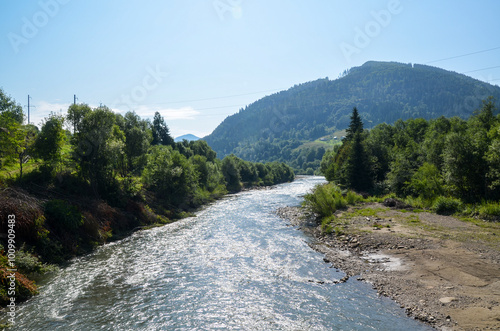 The clear water of Black Cheremosh river flowing through a picturesque valley surrounded by dense forests and rolling hills under a bright blue sky with a few scattered clouds. Carpathian Mountains photo