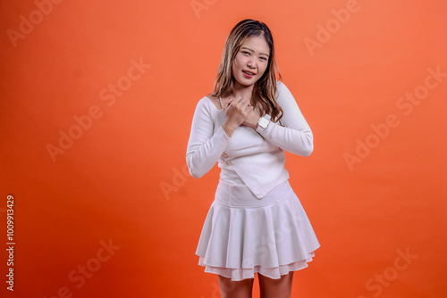 portrait of beautiful young asian woman in white top standing in pain tightness holding chest isolated orange background. for health concept, advertising, science, commercial photo