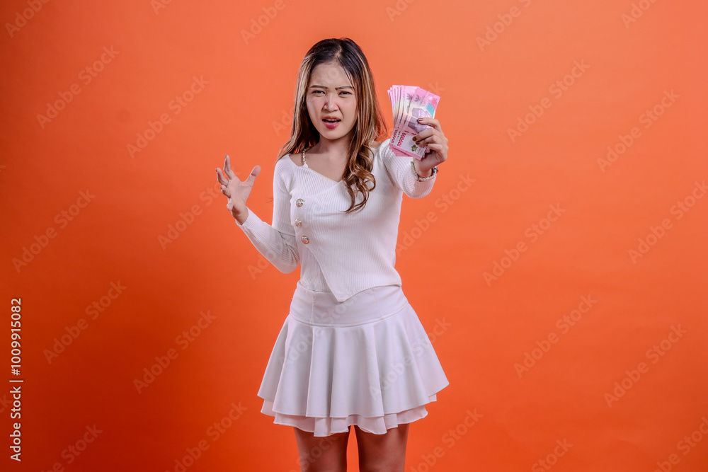 charm of beautiful adult woman white top standing irritated hand holding rupiah cash forward and hand growling isolated orange background. for business concept, competition, finance, reward charm of beautiful adult woman white top standing irritated hand holding rupiah cash forward and hand growling isolated orange background. for business concept, competition, finance, reward