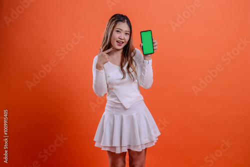 expression of beautiful teenage girl in white top standing cheerfully to camera hand holding mobile phone screen, cellphone while pointing at it isolated orange background. for technology, business