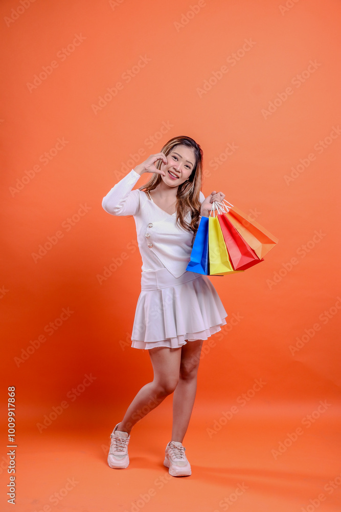 full body charm asian woman white top standing cheerful hands lifting shopping bag while forming love sign like orange background. for fashion concept, advertisement, lifestyle, transaction full body charm asian woman white top standing cheerful hands lifting shopping bag while forming love sign like orange background. for fashion concept, advertisement, lifestyle, transaction