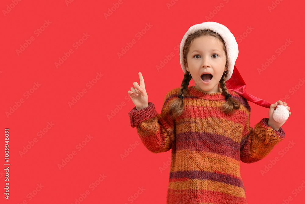 Shocked little girl in Santa hat pointing at something on red background Shocked little girl in Santa hat pointing at something on red background
