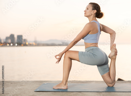 Young slender woman practices yoga on deserted seashore doing asanas, performs physical exercises. During sunset, girl performs Ardha Anjaneyasana photo