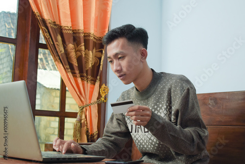 young man shopping online in e commerce, holding credit card and smartphone with laptop om table in livingroom photo