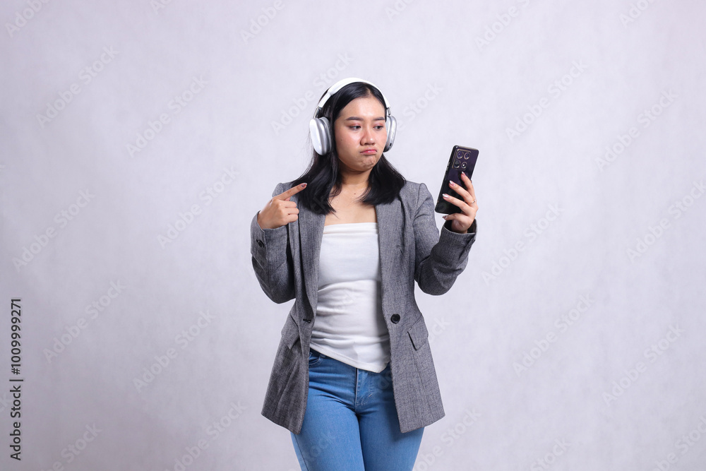 beautiful office lady indonesia pouting at device wearing headphone and holding mobile phone while pointing wearing suit isolated on white background. lifestyle, entertainment, technology concept beautiful office lady indonesia pouting at device wearing headphone and holding mobile phone while pointing wearing suit isolated on white background. lifestyle, entertainment, technology concept