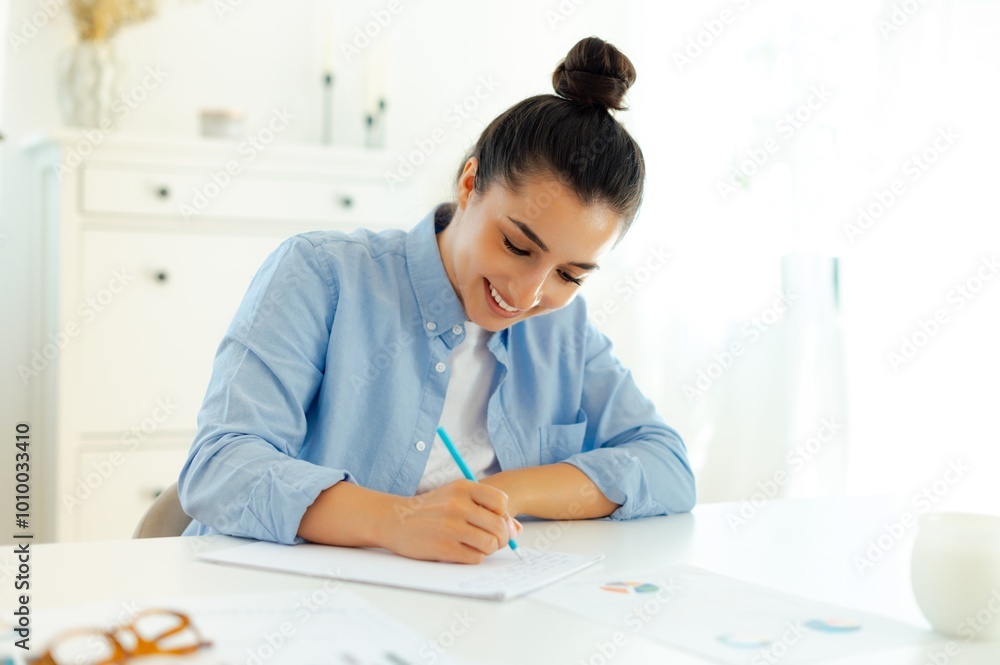 Focused positive caucasian brunette young woman in a blue shirt, sitting at a work desk, writing notes, prepare presentation for colleagues, a female student is preparing for the session, smiling Focused positive caucasian brunette young woman in a blue shirt, sitting at a work desk, writing notes, prepare presentation for colleagues, a female student is preparing for the session, smiling
