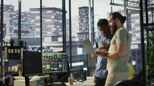 IT employees in office working together using laptop, testing and deploying programs and systems. Developer and colleague checking code on notebook, doing brainstorming in workspace, camera B photo