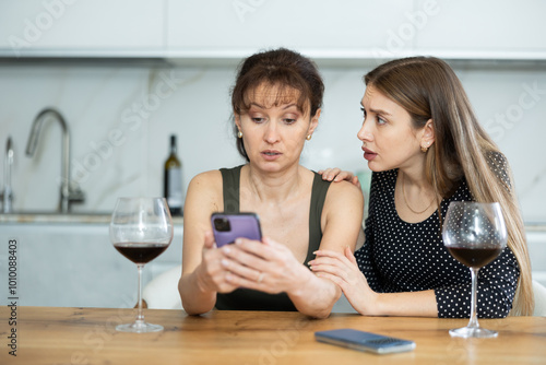 Upset ladies in their middle ages gazing at the phone screen regretfully while drinking a glass of wine photo