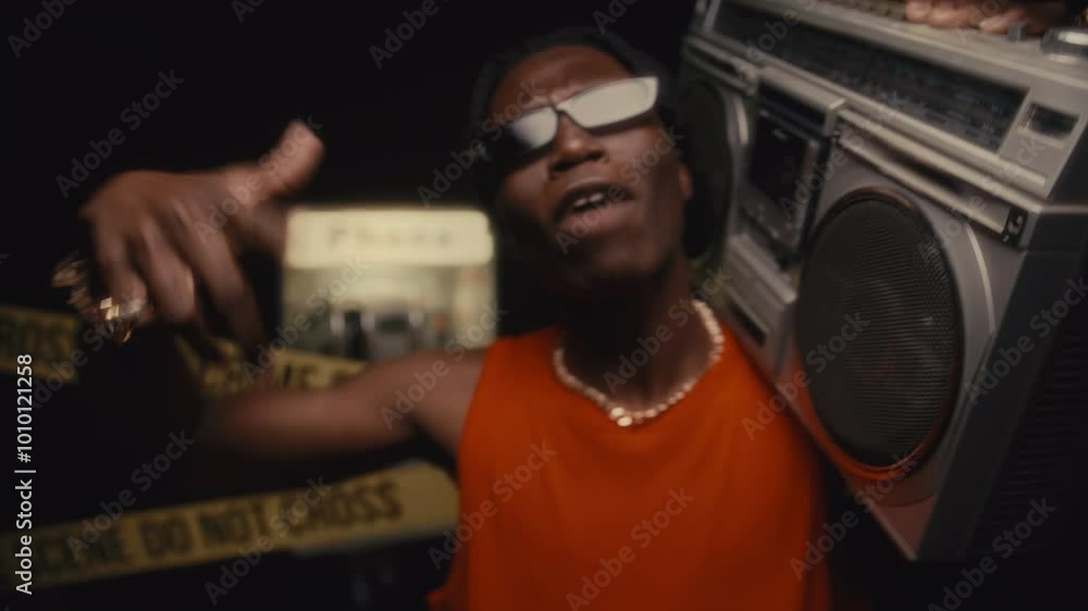 Black hip-hop artist standing at crime scene, holding boombox on his shoulder, rapping into camera with bold hand gestures in front of yellow caution tape, phone booth and dark background Black hip-hop artist standing at crime scene, holding boombox on his shoulder, rapping into camera with bold hand gestures in front of yellow caution tape, phone booth and dark background