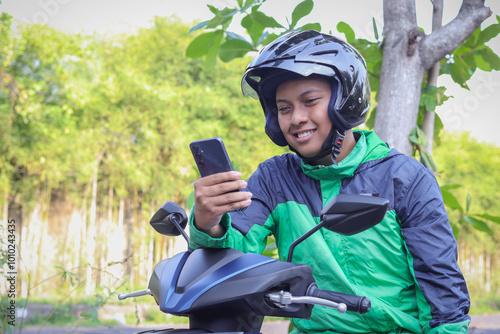 Online taxi rider wearing green jacket and helmet, riding motorcycle, using a mobile phone while on the way to pick up customer. Transportation application concept. photo