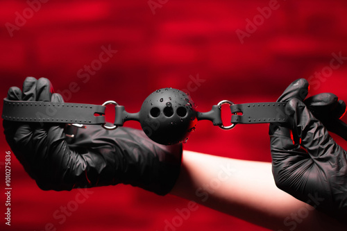 Canvas Print Close-up of female hands in black latex gloves holding a black BDSM gag on a red background