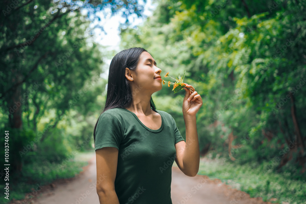 Fototapeta premium An Asian woman is happily relaxing in a green forest garden, smelling flowers and enjoying the fresh air. She takes a deep breath, looks up at the morning sunlight, and feels at peace with nature